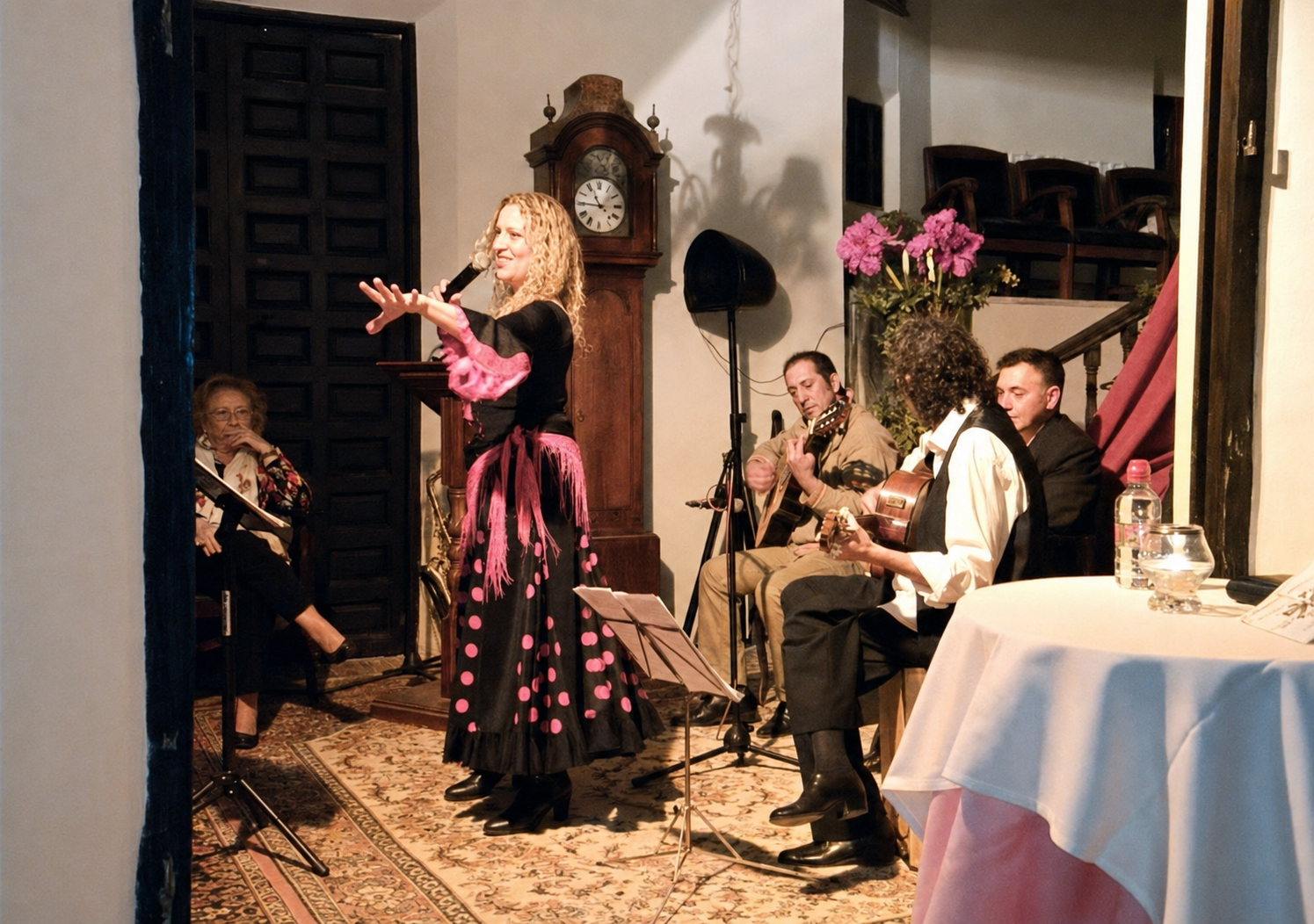 Mona cantando con vestido negro de lunares rosas junto a un cuarteto, con un reloj de pie antiguo al fondo, en la Casa de los Alcaínes