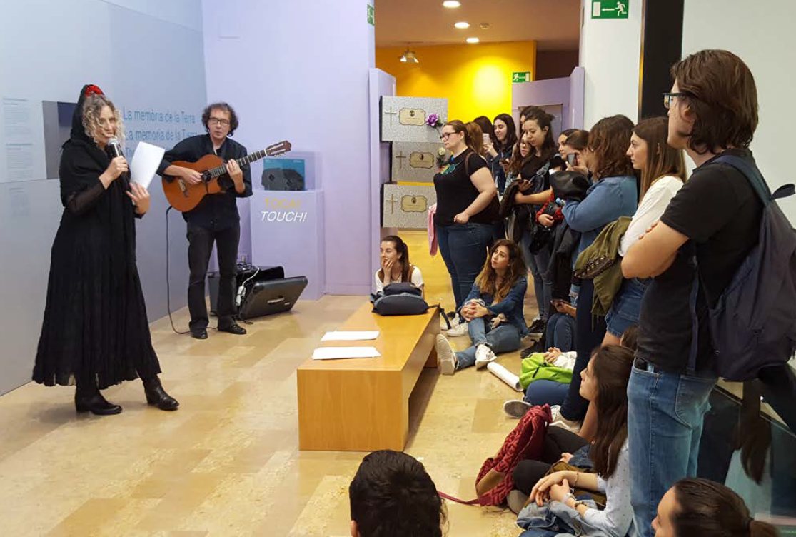 Mona cantando con micrófono y Mike a la guitarra en una sala del Museu de Ciències Naturals de València, rodeados de estudiantes sentados en el suelo y de pie junto a las instalaciones