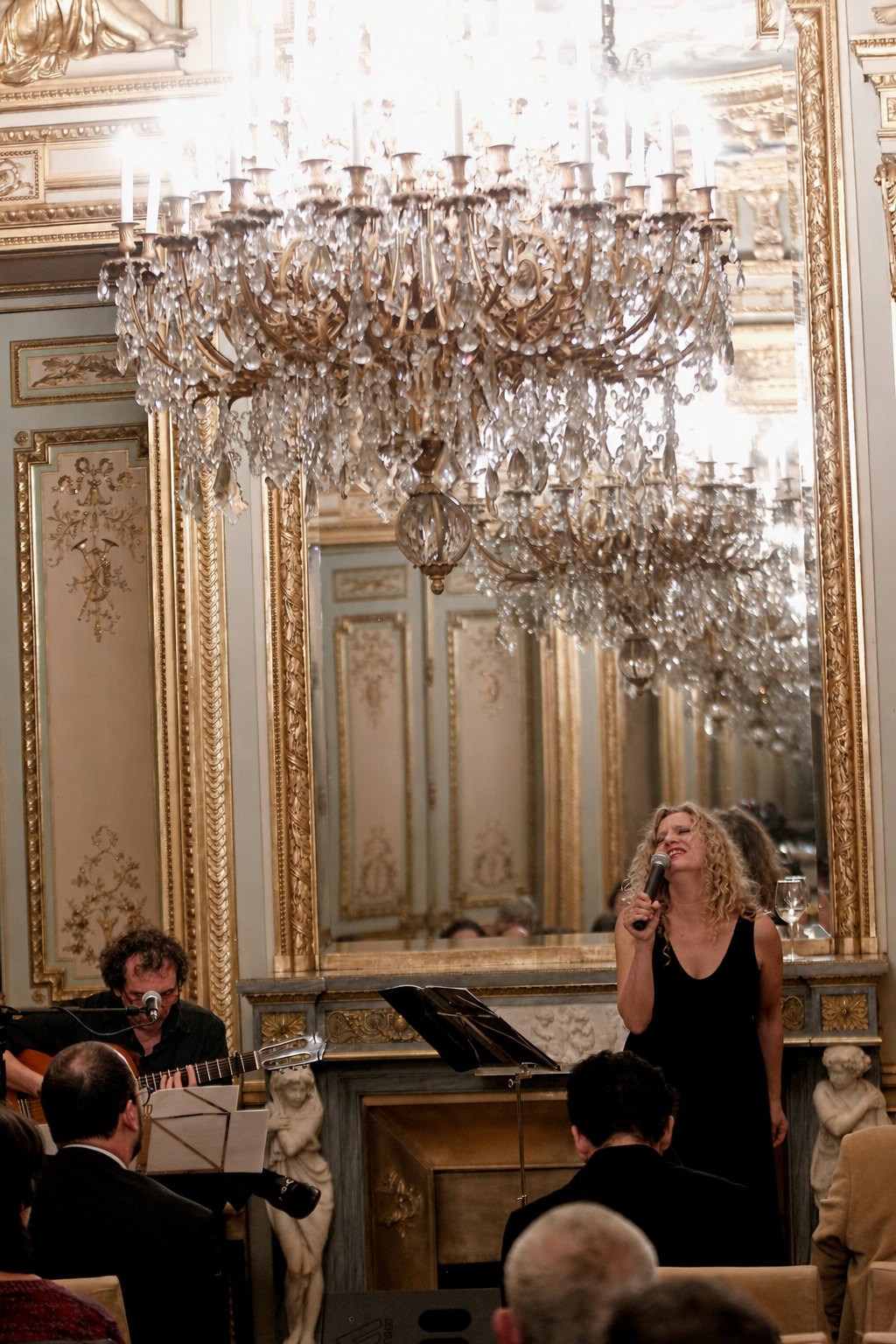 Mike y Mona en concierto: Mona cantando y Mike a la guitarra bajo una lámpara de araña en un salón dorado