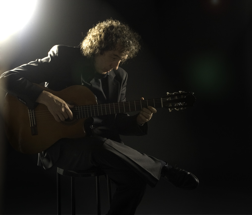 Mike concentrado tocando la guitarra española, sentado en penumbra con luz lateral
