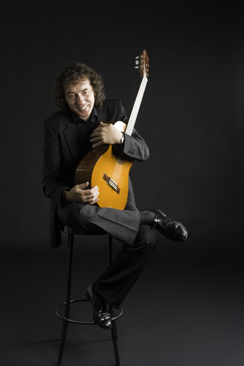Mike sonriendo sentado en un taburete con la guitarra en brazos, retrato de estudio
